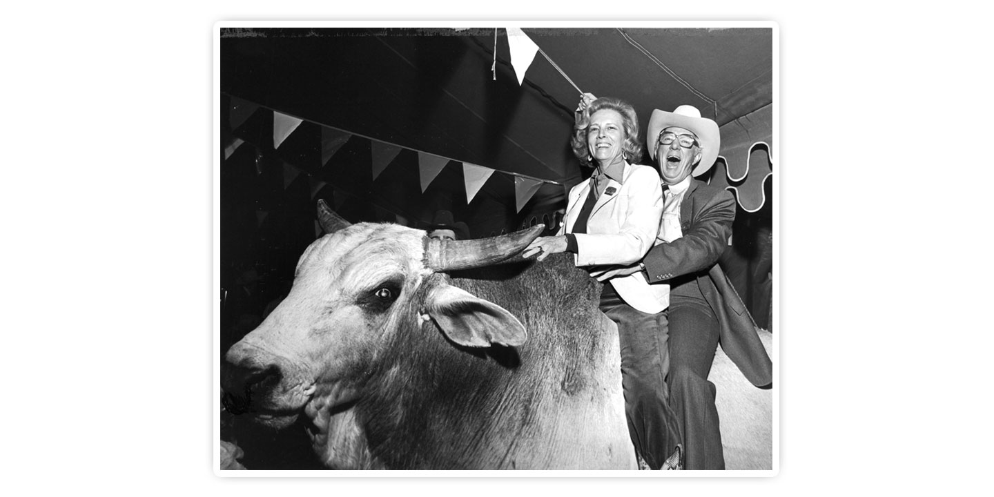 Mr. John S. Justin, Jr. and Mrs. Jane Chilton Justin pose on a Bucking Bull, smiling and laughing, holding each other.